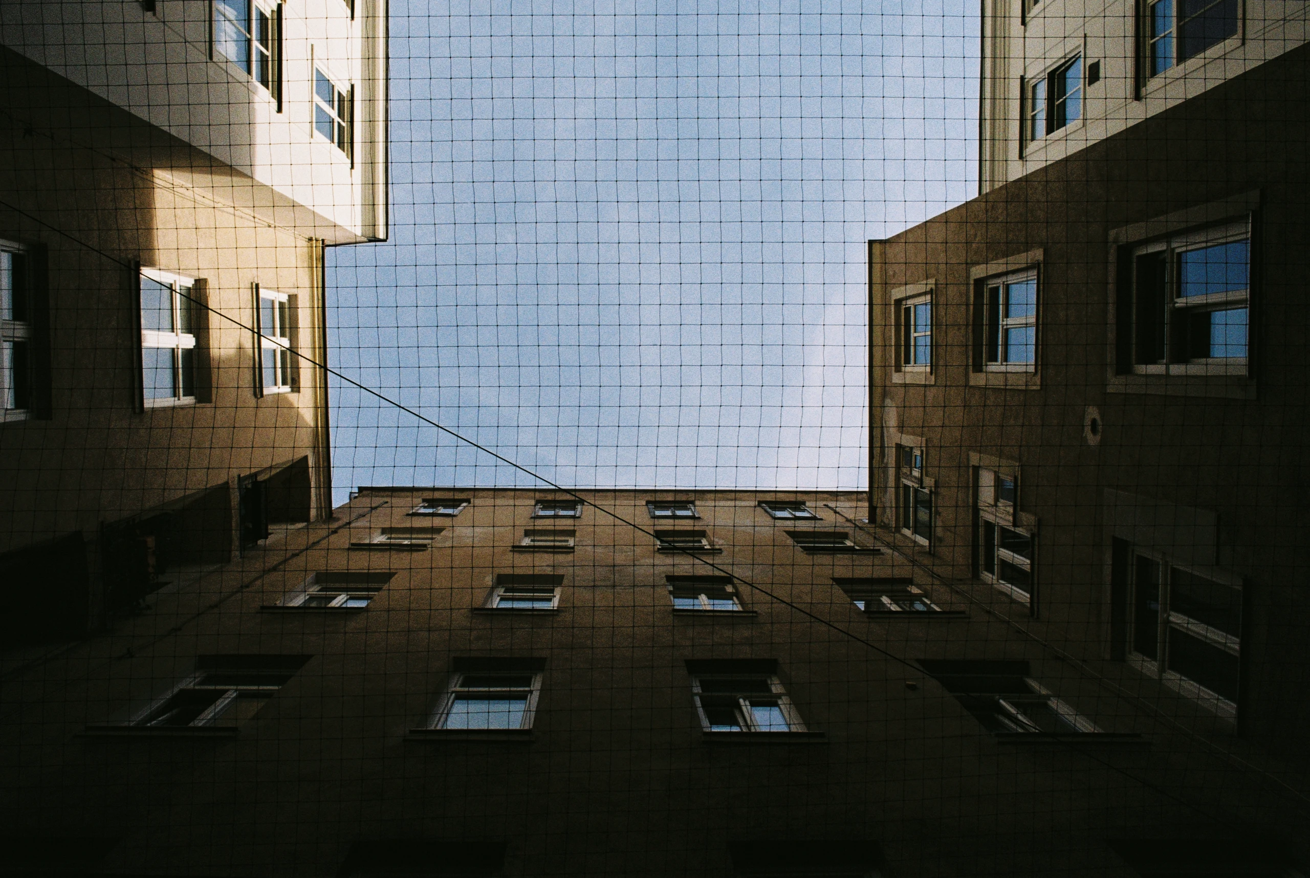 Aufnahme aus der Perspektive eines Netzes, das den Blick auf einen Innenhof mit mehreren Gebäuden und Fenstern blockiert. Die Gebäude sind in gedämpften Braun- und Grautönen gehalten. Der Himmel ist blau und durchbrochen von einem Netz aus dunklen Linien. Die Fenster reflektieren das Licht und erzeugen helle Kontraste. Die Komposition ist symmetrisch, mit den Gebäuden links und rechts und einem zentralen Himmel. Die Lichtquelle ist oben, was zu Schatten auf den Wänden führt.