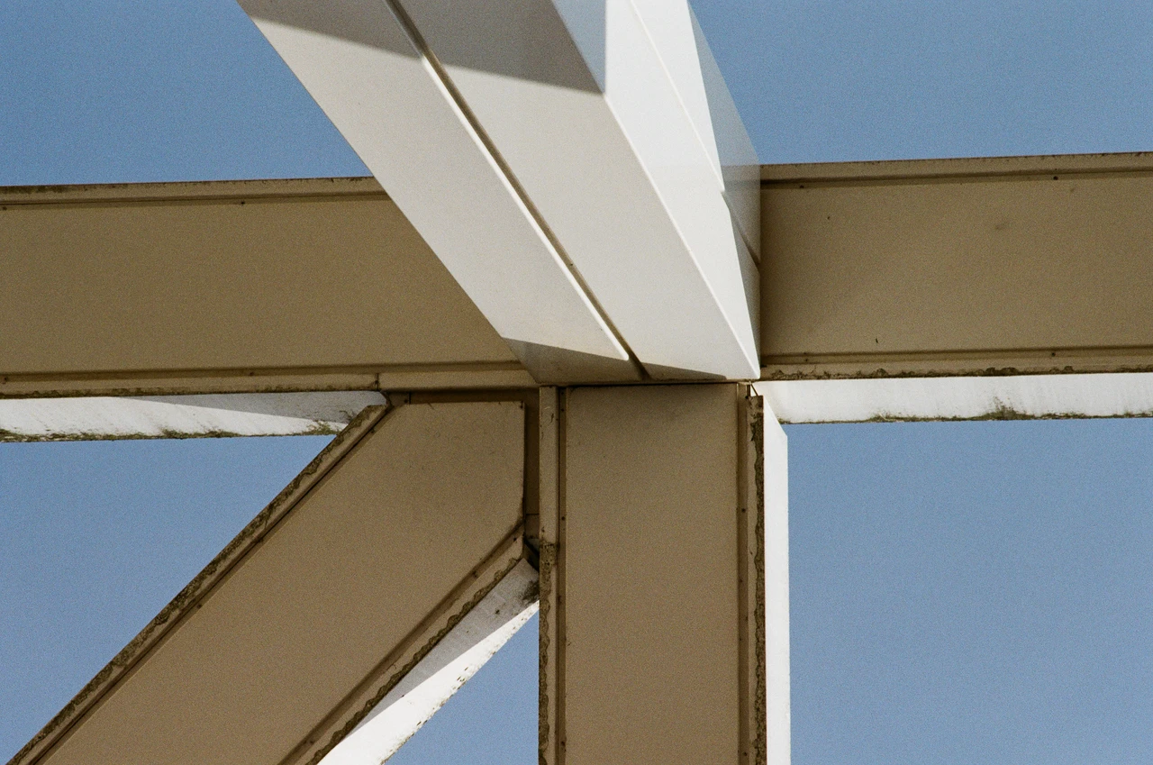 Nahaufnahme eines metallischen Stahlrahmens, der sich im Winkel von 90 Grad kreuzt. Der Rahmen ist in einem hellen, beigefarbenen Ton lackiert, mit sichtbaren Schmutz- und Moosablagerungen an den Kanten. Der Hintergrund besteht aus einem klaren, blauen Himmel. Die Komposition ist asymmetrisch, mit diagonalen Linien, die sich im Zentrum treffen. Die Beleuchtung ist natürlich und erzeugt harte Schatten auf den Oberflächen.