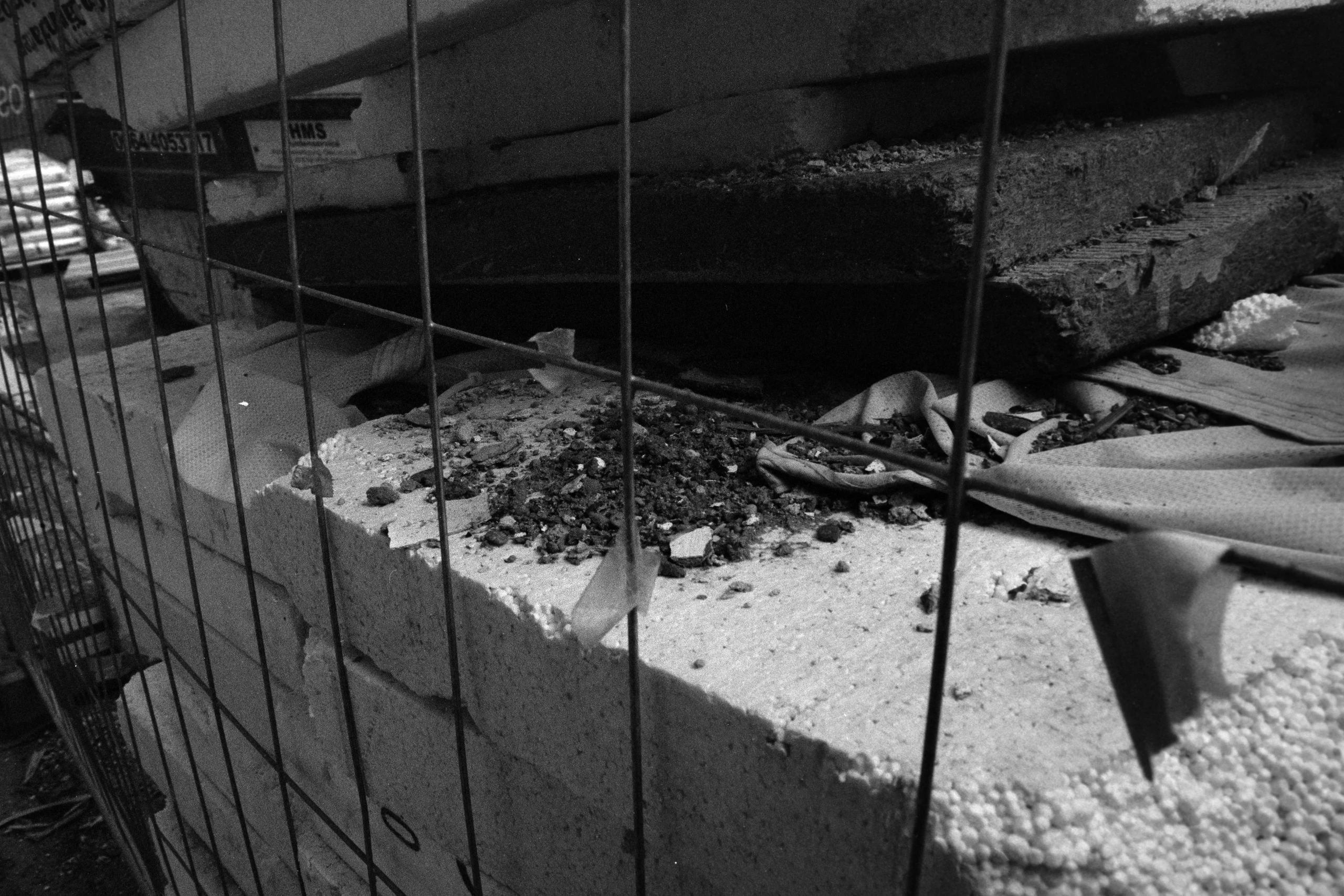 Schwarz-Weiß-Aufnahme eines Baustellenbereichs, der durch ein Stahlgitter im Vordergrund eingerahmt ist. Im Hintergrund sind zerbrochene Ziegel und Styroporplatten zu sehen, die auf einem Boden aus Schutt und Erde liegen. Die Komposition ist asymmetrisch, mit starken Kontrasten zwischen Licht und Schatten.
