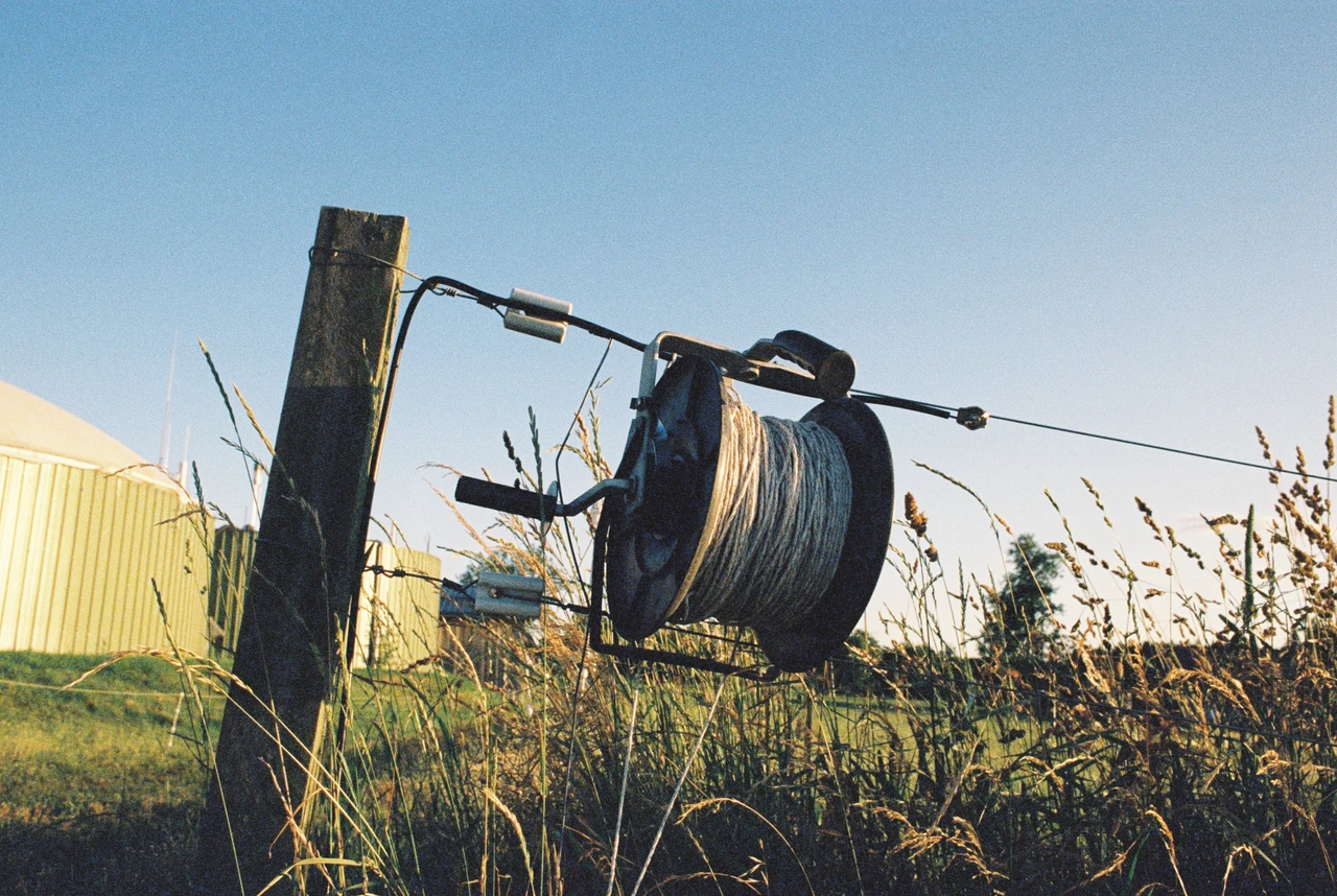 Ein Drahtseilwinder aus Metall, befestigt an einem Holzpfosten, hängt über einem Feld mit hohem Gras. Im Hintergrund ist ein gelbes Wellblechgebäude zu sehen. Die Szene wird von hellem Tageslicht beleuchtet, das Schatten auf den Boden wirft.