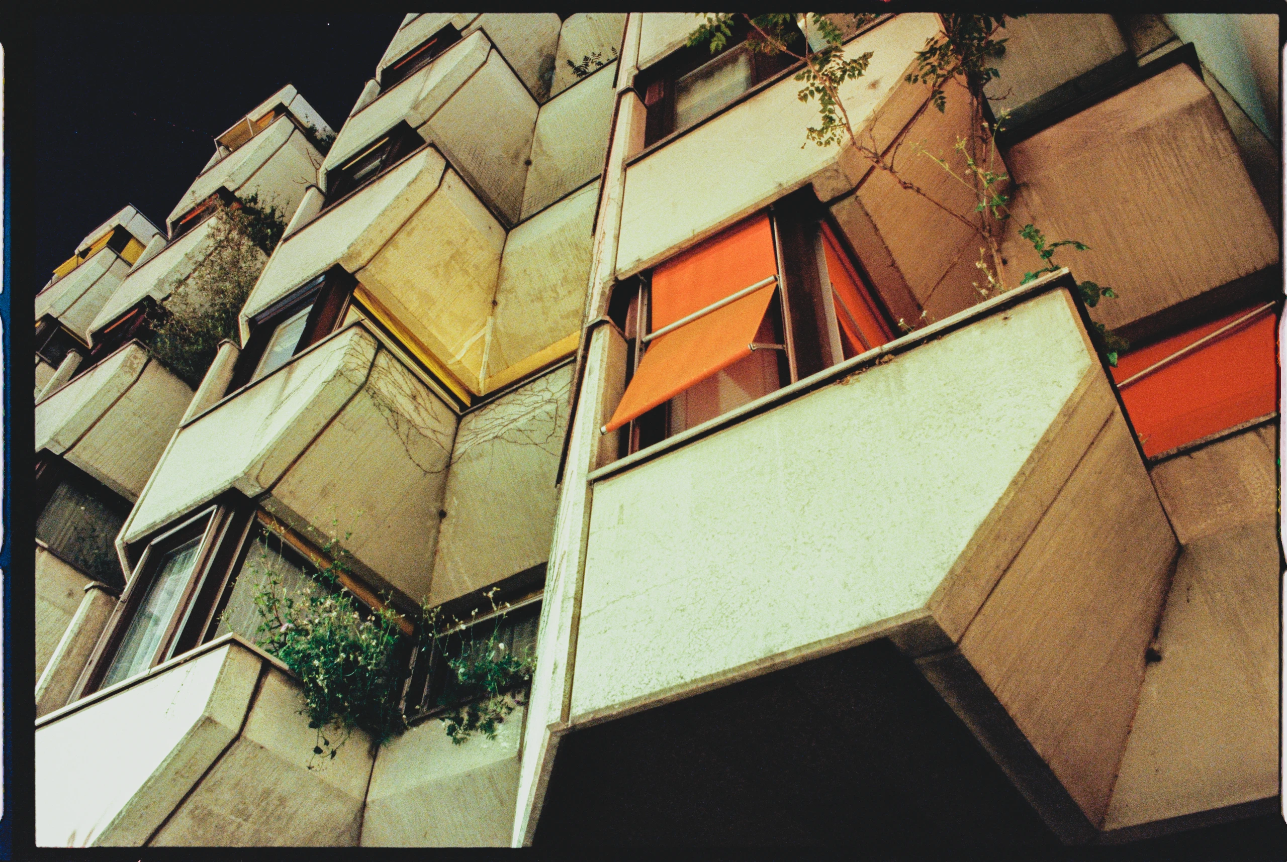 Nahaufnahme eines Wohnhauses bei Nacht, fotografiert aus einer schrägen Perspektive. Die Fassade besteht aus betonartigen, geometrischen Elementen in beige- und grautönen. Mehrere Balkone sind mit orangefarbenen Markisen und grünen Pflanzen versehen. Der Hintergrund ist dunkel, was die strukturellen Linien und die Textur der Oberflächen hervorhebt.