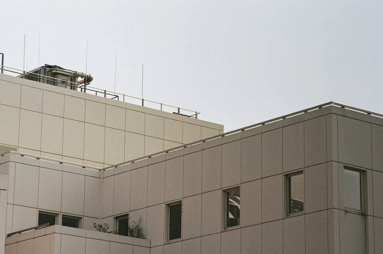 Nahansicht der oberen Gebäudewand aus hellen, quadratischen Paneelen. Im oberen Bereich sind Dachantennen und ein technisches Gerät auf einem Geländer zu sehen. Rechts befindet sich ein Eckabschnitt mit mehreren Fenstern. Der Himmel ist bedeckt und erzeugt diffuses Licht. Einige Pflanzen wachsen an der Wand.