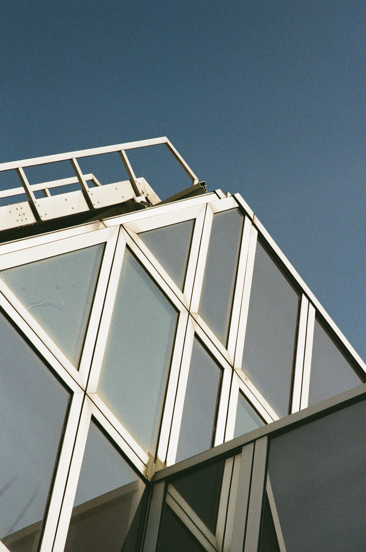 Nahaufnahme der oberen Ecke eines modernen Gebäudes mit geometrischem Glas- und Metallrahmen. Der Himmel ist klar und blau. Die Sonne beleuchtet die weißen Rahmen von oben, während die Glasflächen den Himmel reflektieren. Im oberen linken Bereich ist eine Metallbrüstung sichtbar. Die Komposition ist asymmetrisch und zeigt eine diagonale Linie, die vom unteren linken zum oberen rechten Bildrand führt.