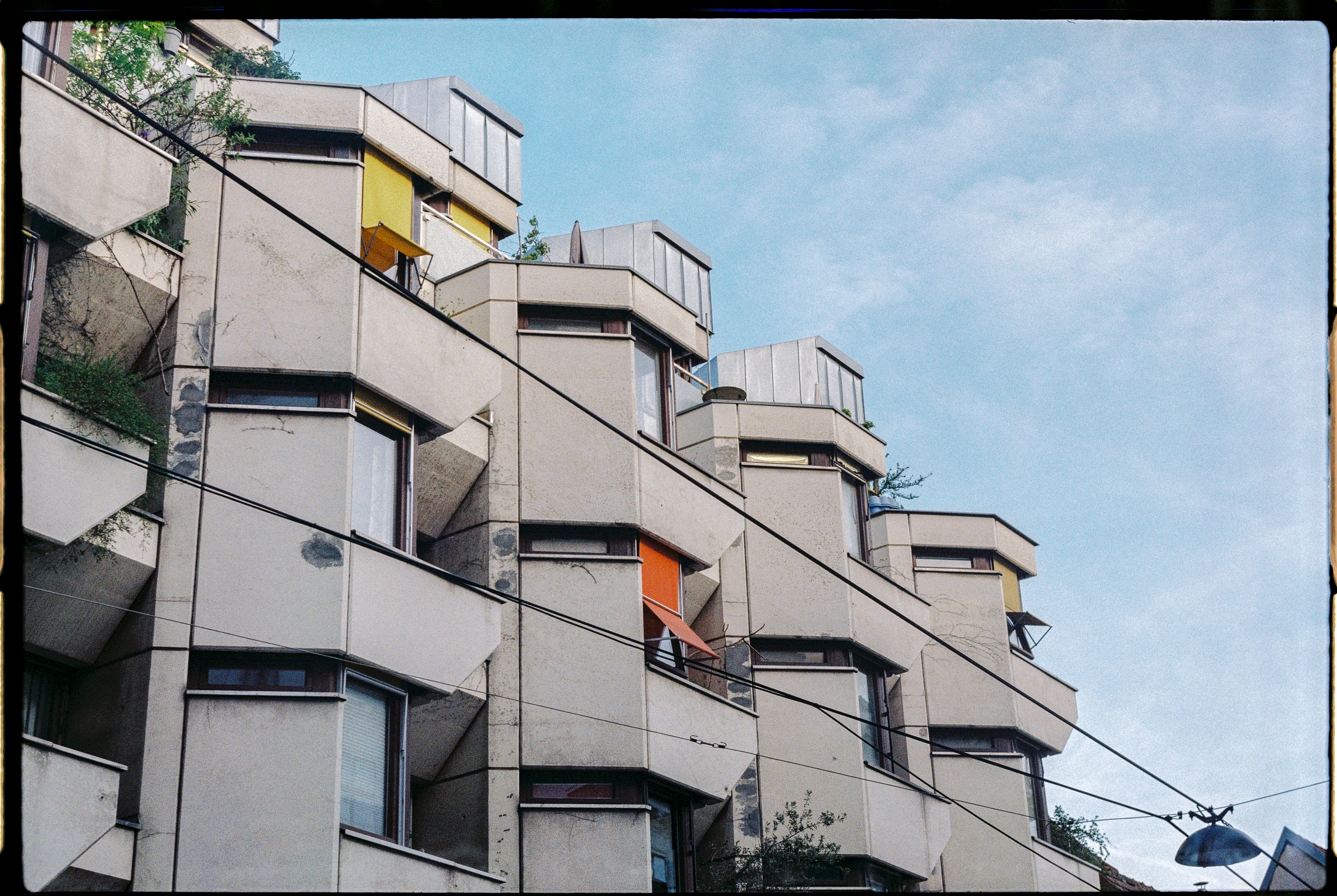 Niedriger Blickwinkel auf ein mehrstöckiges Wohnhaus mit betonierten, geometrisch angeordneten Balkonen. Mehrere Balkone sind mit gelben und orangefarbenen Markisen ausgestattet. Dunkle Stromkabel verlaufen diagonal über das Bild. Der Himmel ist hellblau mit weißen Wolken. An einigen Balkonen wachsen Pflanzen. Die Fassade zeigt Spuren von Verwitterung und Schmutz.