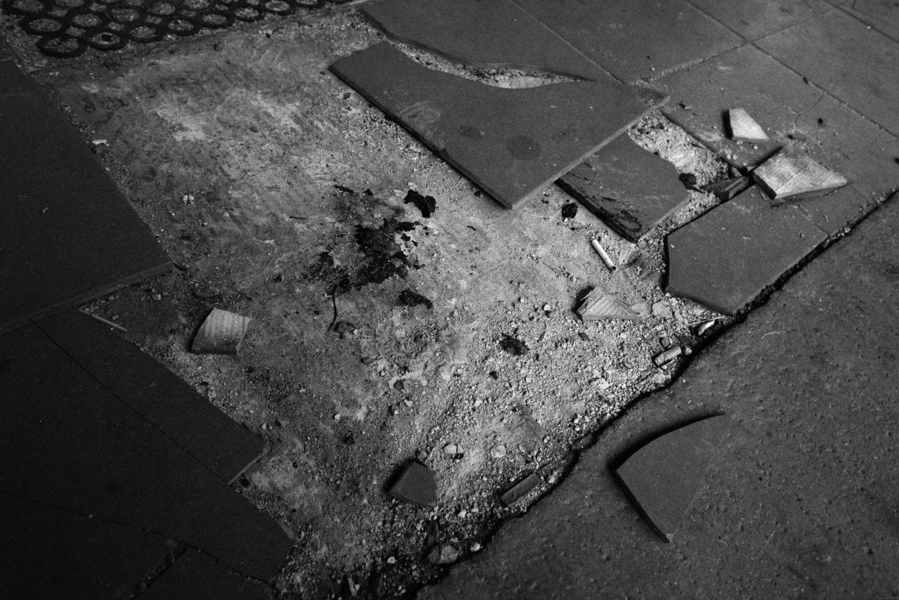 Schwarz-Weiß-Aufnahme eines zerstörten Gehwegabschnitts. Im Zentrum befindet sich ein unregelmäßiger Bereich aus Erde und Schutt, umgeben von intakten und zerbrochenen Fliesen. Die Komposition ist diagonal ausgerichtet, mit scharfen Schatten und hohen Kontrasten. Sichtbare Objekte: Zerbrochene Fliesen, Erde, Asche, Zigarettenstummel, ein abgebrochener Zylinder. Materialien: Beton, Ziegel, Erde, Metall. Die Fliesen sind in verschiedenen Größen und Formen zerbrochen und liegen unregelmäßig verteilt.
